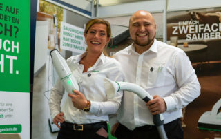 Lachendes Paar in weißen Hemden mit Staubsaugerschlauch an einem Messestand – Fotoshooting auf der „Bleib fit“ im M.T.C. München