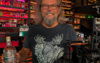Michael J. Müller hält eine Ginflasche in der Bar des Gin City Munich, daneben die Kamera – Dreh für den Imagefilm