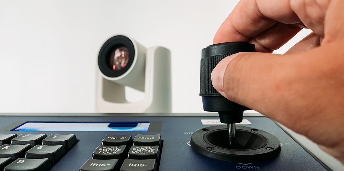 Hand am Joystick des Kamera-Controllers, im Hintergrund die PTZ-Kamera – Fernsteuerung im Livestream